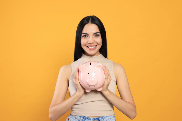 Happy young woman with piggy bank on orange background - Фото, зображення