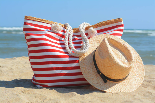 Stylish striped bag with straw hat on sandy beach near sea - Fotó, kép