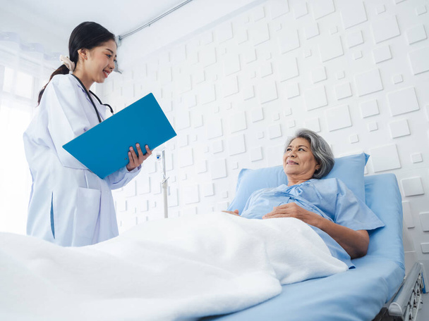Uśmiechnij się Azjatka seniorka siwe włosy pacjenta leżącego na łóżku, dochodzi do siebie podczas gdy młode kobiety lekarze odwiedzają, wyjaśniając objawy i notatki z badań i dokumentów medycznych opieki zdrowotnej w pokoju szpitalnym.  - Zdjęcie, obraz
