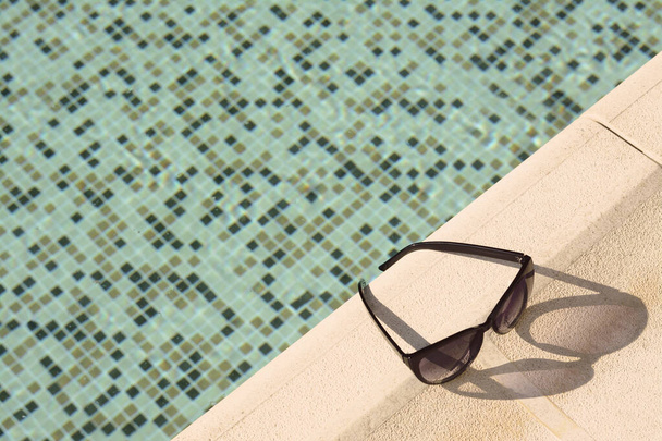 Stylish sunglasses near outdoor swimming pool on sunny day, above view. Space for text - Φωτογραφία, εικόνα