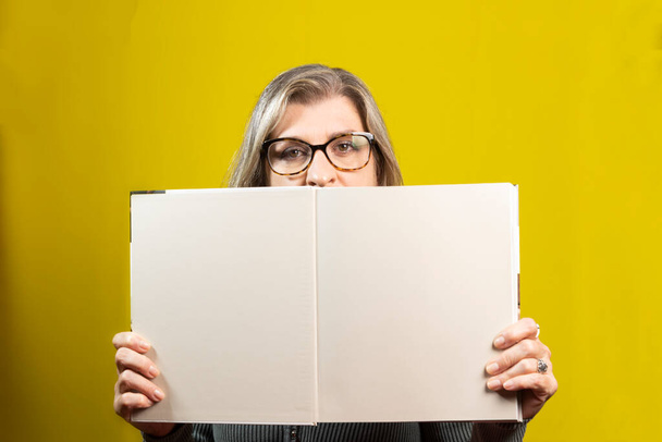 Žena středního věku ukazuje na kameru otevřenou knihu s prázdnými stránkami na žlutém pozadí. Koncept vzdělávacího životního stylu. - Fotografie, Obrázek