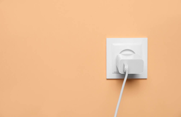 Charger adapter plugged into power socket on pale orange wall, space for text. Electrical supply - Photo, Image