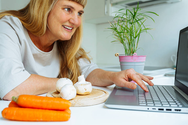 Жінка середнього віку з довгим світлим волоссям кухарів на кухні і дивиться на екран ноутбука з цікавістю. Жінка готує вегетаріанську страву, овочі на столі. Готувати овочі вдома на кухні згідно з рецептом з Інтернету. - Фото, зображення