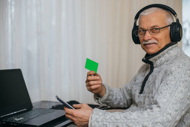 Senior man working at laptop at home. Old man enters credit card number into phone. Loans for pensioners. Opening retirement accounts. elderly man shows credit card. mockup. Clipping path - Photo, Image