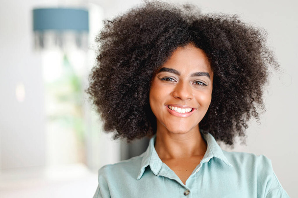 Headshot of beautiful smiling serene African-American young woman in casual shirt resting on the sofa at cozy apartment, charming multiracial girl looking at the camera with wide smile - Foto, imagen
