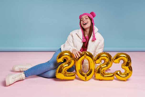 Cheerful young woman in winter coat holding gold colored numbers while sitting against blue background - Фото, зображення