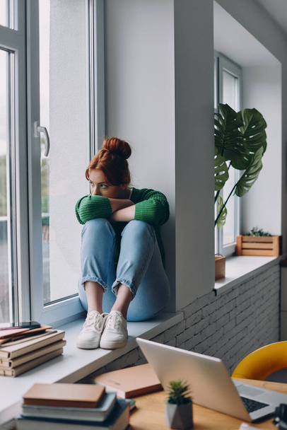 Young redhead woman looking depressed while sitting on the window sill at home - 写真・画像