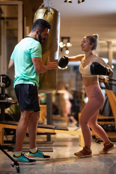 Fit girl with boxing gloves hitting a punching bag. Male fitness instructor with a female client practicing kickbox in a fitness studio. - Фото, изображение