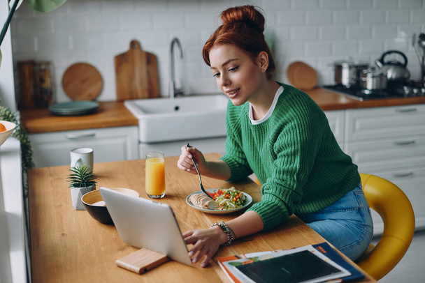 Attractive young woman having lunch and using digital tablet while sitting at the kitchen counter - Valokuva, kuva