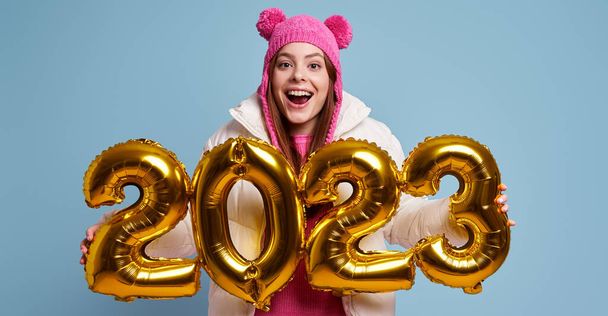 Excited young woman in winter coat holding gold colored numbers against blue background - Фото, зображення