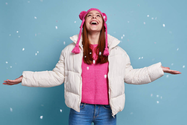 Boldog nő télikabátban tartja karját kinyújtva, miközben hó esik a kék háttér - Fotó, kép