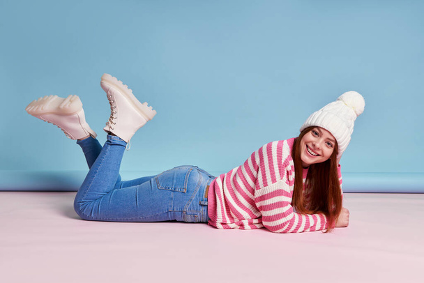 Happy young woman in winter clothing looking at camera while lying against blue background - Foto, Imagen