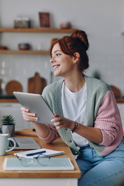 Glückliche junge Frau nutzt digitales Tablet, während sie zu Hause am Küchentisch sitzt - Foto, Bild