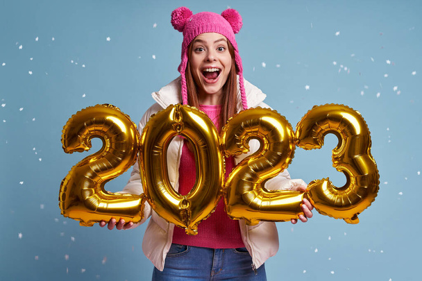 Beautiful woman in winter coat holding gold colored numbers and smiling against blue background - Foto, Imagem