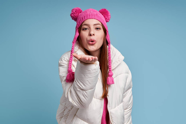 Beautiful young woman in winter coat blowing a kiss while standing against blue background - Foto, Imagem