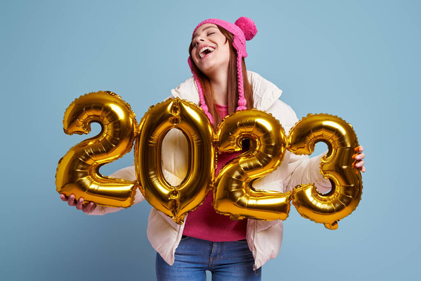 Happy young woman in winter coat holding gold colored numbers against blue background - Foto, afbeelding
