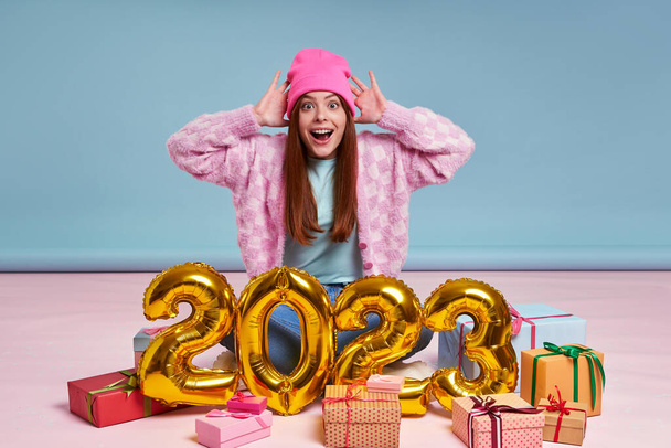Surprised young woman sitting near the gift boxes and gold colored numbers against blue background - Foto, Imagen