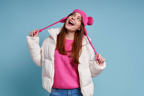 Beautiful young woman in winter coat and funky hat looking up and smiling against blue background - Foto, afbeelding