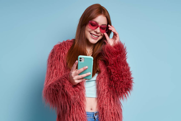 Fashionable young woman in fluffy coat using smart phone against blue background - Fotoğraf, Görsel