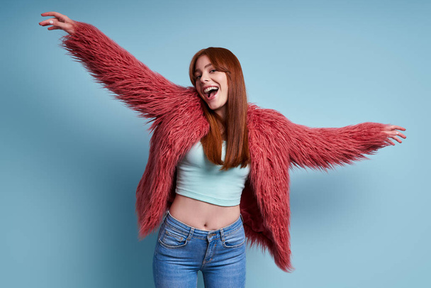 Excited young woman in fluffy coat dancing against blue background - Foto, Imagem