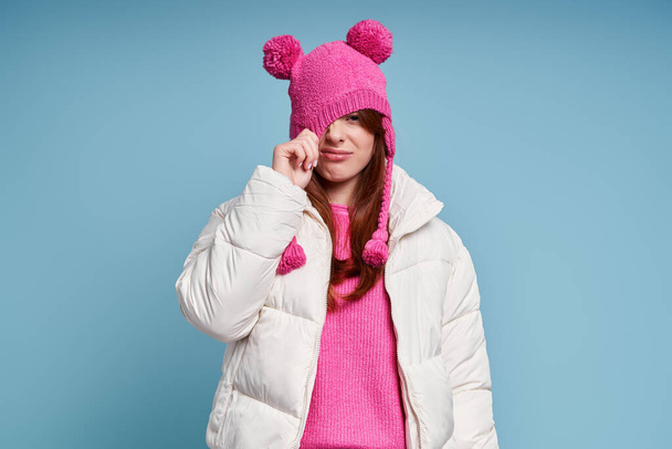Displeased young woman in winter coat pulling her funky hat while standing against blue background - Foto, imagen