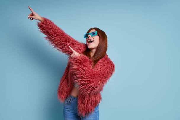 Happy young woman in fluffy coat pointing away while standing against blue background - Fotoğraf, Görsel