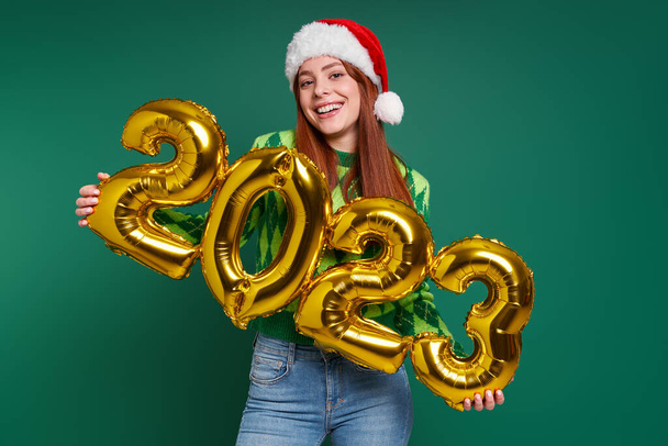Happy woman in Christmas hat holding gold colored numbers against green background - Foto, imagen
