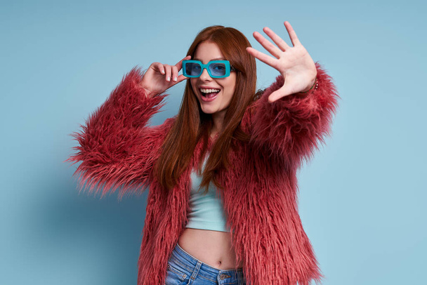 Fashionable young woman in fluffy coat smiling and gesturing against blue background - Valokuva, kuva