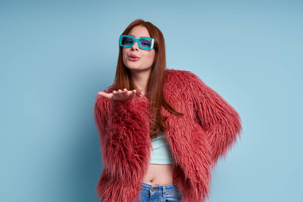 Happy young woman in fluffy coat blowing a kiss while standing against blue background - Fotografie, Obrázek