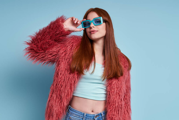 Fashionable young woman in fluffy coat adjusting her eyeglasses against blue background - Foto, imagen