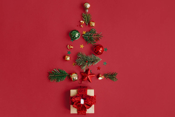 Merry Christmas! Stylish christmas tree made of fir branches, baubles, gold confetti and gift on red background, festive flat lay. Creative idea. Winter holidays, modern banner - Фото, зображення