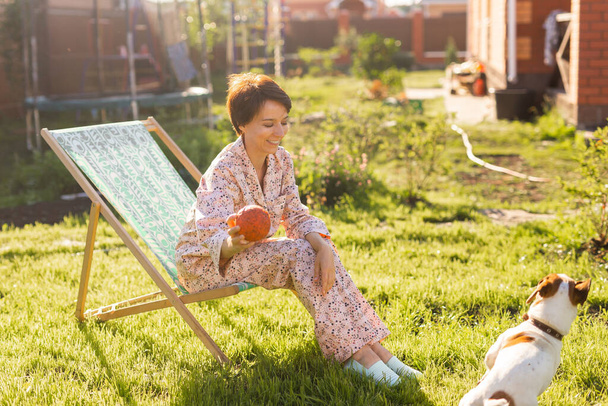 Jonge vrouw in pyjama rust in stoel op een groen gazon op zonnige zomerdag - dorps- en landleven concept - Foto, afbeelding