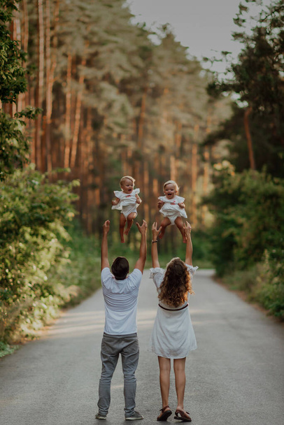 a family walks in the middle of nature with two children, twin sisters; dad and mom throw their little sisters up; the family is standing on the asphalt road in the middle of the forest - Zdjęcie, obraz