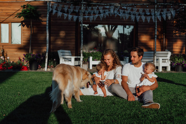 rodina prochází uprostřed přírody se dvěma dětmi, dvojčaty, rodiče si hrají se svými dětmi v blízkosti domu na zeleném trávníku a jejich pes je vedle nich - Fotografie, Obrázek