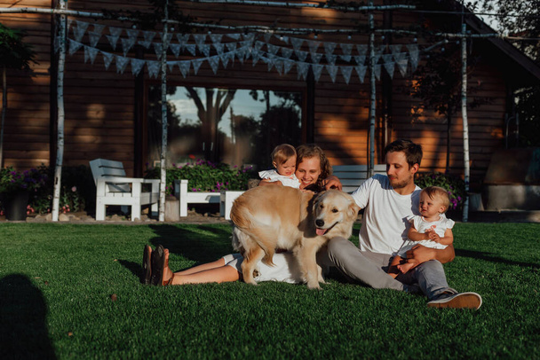 rodzina spaceruje po środku natury z dwójką dzieci, bliźniaczkami; rodzice bawią się z dziećmi w pobliżu domu na zielonym trawniku, a ich pies jest obok nich - Zdjęcie, obraz