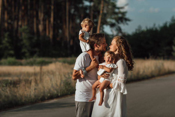una famiglia cammina in mezzo alla natura con due bambini, sorelle gemelle; i genitori tengono i bambini tra le braccia; papà bacia la mamma - Foto, immagini