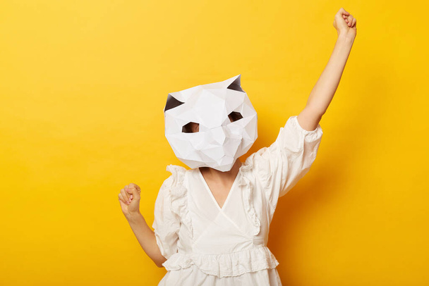 Indoor shot of extremely happy unrecognizable woman wearing dress and paper cat mask posing isolated over yellow background, raised her arms, winning, celebrating. - Foto, Imagen