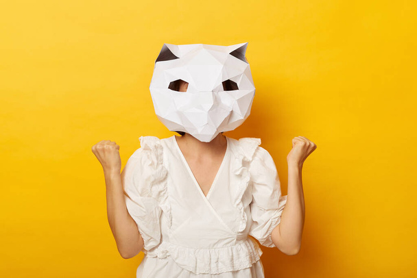 Indoor shot of anonymous woman wearing dress and paper cat mask, showing her clenched fist, celebrating victory, posing isolated over yellow background. - Valokuva, kuva