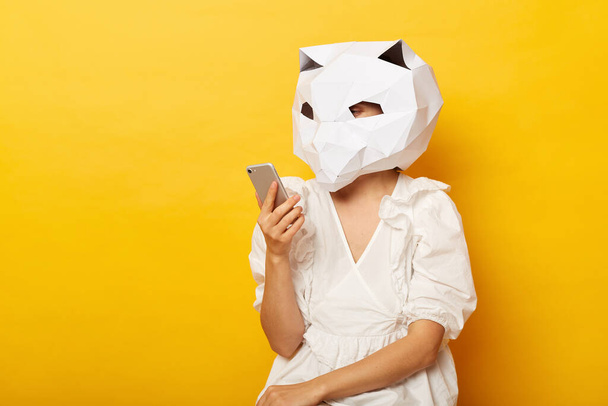 Indoor shot of woman wearing dress and paper cat mask posing isolated over yellow background, holding mobile phone, using smart phone for reading news in Internet. - Photo, image