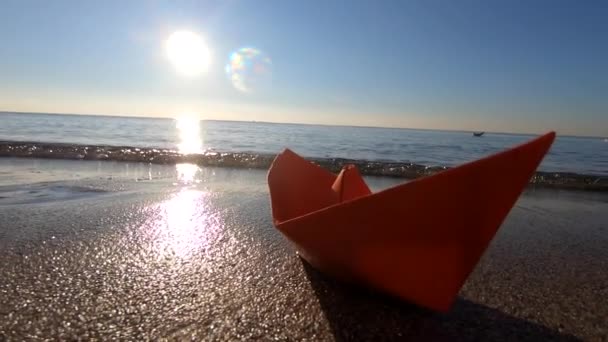 Egy kis papír piros hajó a homokos tengerparton, közel a tenger hullámai egy napsütéses napon. Kilátás alulról közelről. Koncepció, jel, az utazás szimbóluma, turizmus, álmok, célok, nyaralás, kikapcsolódás - Felvétel, videó