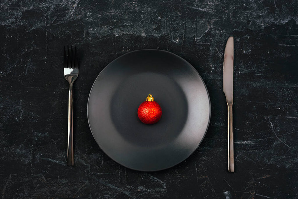 Weihnachtstisch mit roter Weihnachtskugel auf leerem schwarzen Teller und Besteck auf schwarzem Hintergrund. Draufsicht, flache Lage. - Foto, Bild