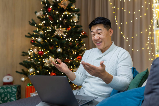 Veselé Vánoce muž dělat videohovor pomocí notebooku sedí na pohovce v obývacím pokoji doma, asijský muž pozdravuje přátele on-line šťastný nový rok s úsměvem a šťastně mluví. - Fotografie, Obrázek