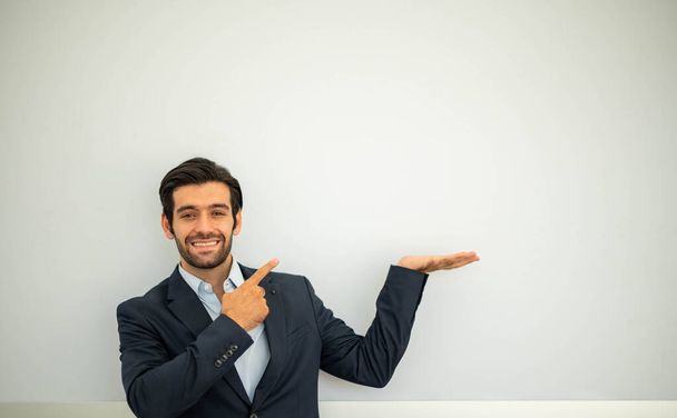 Portrait d'un jeune homme d'affaires caucasien souriant portant un costume sombre et présentant l'espace de copie sur ses paumes et pointant vers le mur de béton. - Photo, image