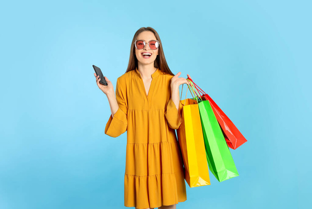 Happy young woman with shopping bags and smartphone on light blue background. Big sale - Foto, Bild