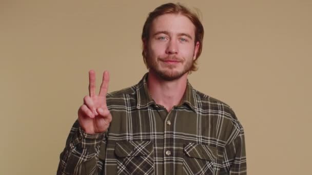 Bearded man in checkered shirt showing victory sign, hoping for success and win, doing peace gesture, smiling with kind optimistic expression. Young adult guy boy isolated on beige studio background - Footage, Video