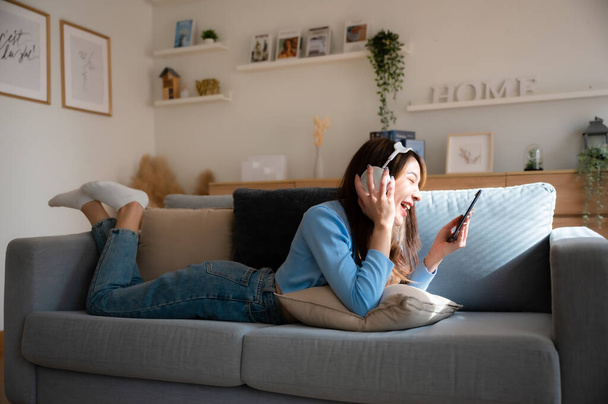 Felice giovane donna asiatica rilassante a casa. Sorriso femminile seduto sul divano e con lo smartphone in mano. Ragazza che usa la videochiamata ad un amico. Ascolto della musica - Foto, immagini