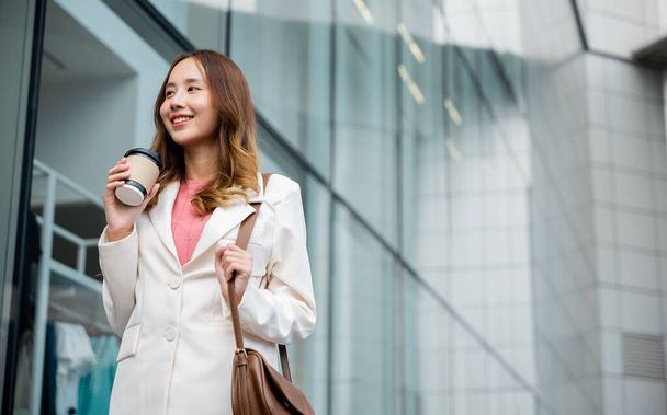 Happy business woman smiling hold paper cup of hot drink outdoor walking on street, Asian businesswoman holding coffee cup takeaway going to work she walking near her office building outside - Zdjęcie, obraz