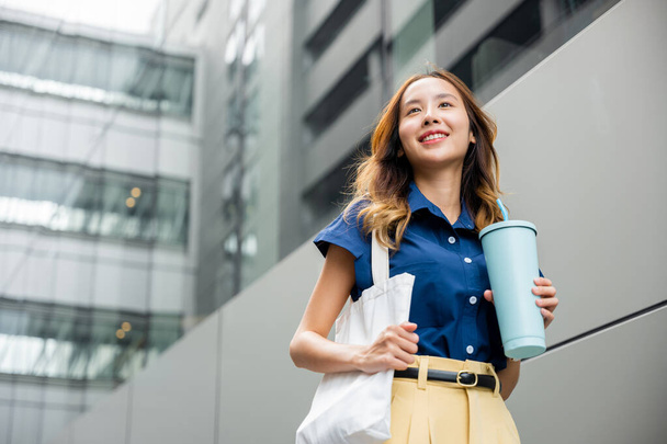 Asian beautiful business woman confident smiling with cloth bag holding steel thermos tumbler mug water glass she walking outdoors on street near modern building office, Happy female - Foto, afbeelding