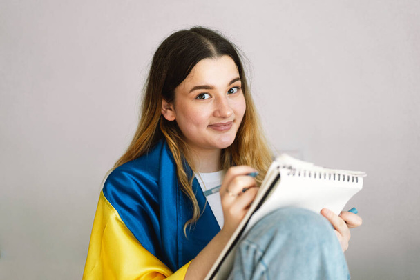 A Ukrainian teenage girl is wrapped in the Ukrainian flag and is drawing or doing homework in the bedroom. A teenager studies at home. The concept of studies during the war in Ukraine. - 写真・画像
