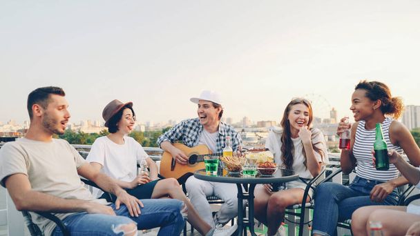 Handsome young man is playing the guitar while his cheerful friends are singing, drinking and laughing sitting at table on rooftop. Music, fun and summertime concept. - Foto, afbeelding